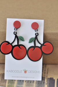 Pair of glossy red cherry drop earrings with black stems and green leaves on Kaboodle card