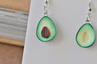 Close-up of two avocado clay earrings showing green skin, cream flesh and brown pit detail