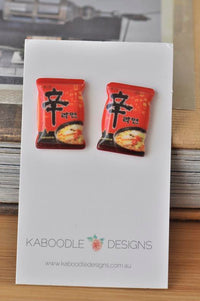 Two resin stud earrings shaped like red Shin Ramyun instant noodle packets on a card