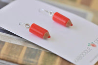 Close-up angled view of two red resin pencil earrings resting on backing card