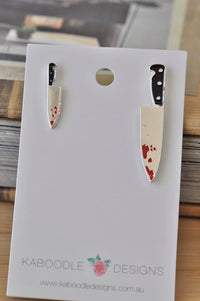 Mismatched silver knife earrings with black handles and red blood splatter on white display card