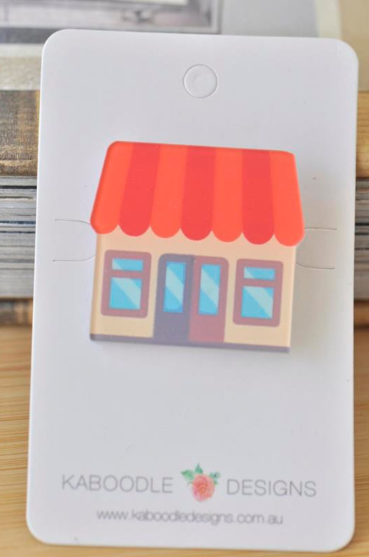 Acrylic brooch shaped like a candy shop house with a red striped awning and blue windows