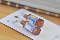 Close-up angled view of two amber resin bottle earrings on display card, showing glossy finish