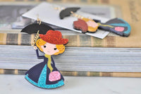 Pair of umbrella and nanny drop earrings laid on books beside earring card