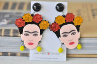 Pair of Frida Kahlo drop earrings on branded card, front view against stacked books