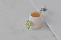 Close-up of white teacup earring with amber tea and green Lipton-style tea bag tag
