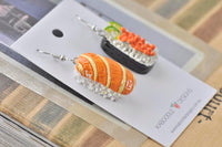 Sushi and sashimi ceramic earrings on card against wooden background, angled view