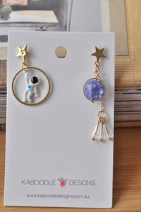 Mismatched astronaut and galaxy stone drop earrings on white display card with gold star studs