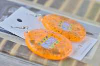 Orange Tamagotchi drop earrings on card resting on wooden surface, angled view