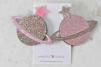 Flat lay of glitter Saturn earrings on branded card, one pink planet and one multi-glitter planet with star studs