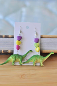 Pair of brontosaurus dinosaur earrings with heart bead dangles on display card, toy dinosaurs beside them