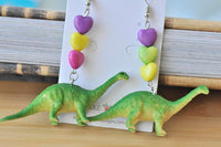 Front view of both brontosaurus dangle earrings with purple, yellow and pink heart beads, toy dinosaurs in foreground