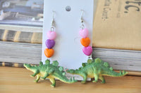 Pair of stegosaurus earrings with heart beads laid flat on jewellery card against books