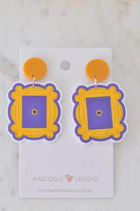 Acrylic door frame earrings with yellow scrollwork frame and purple centre, on orange studs, shown on backing card