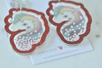 Angled close-up of two unicorn earrings showing pearl beads settled inside the glittery red outline