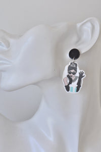 Close-up of single earring worn on ear, showing the printed figure in sunglasses waving with a gloved hand