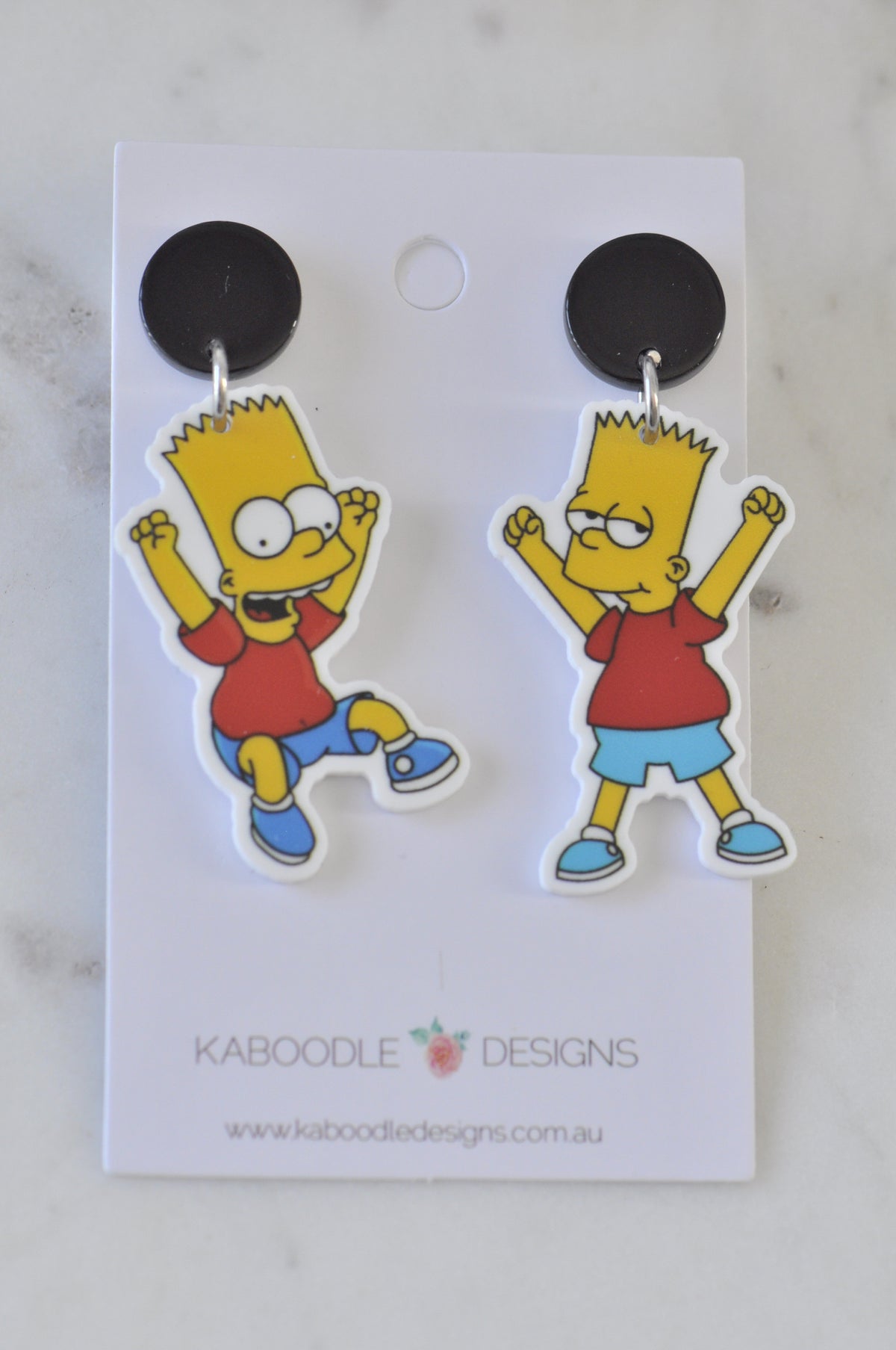 Pair of Bart Simpson dangle earrings on black round studs, displayed on a card against a marble background