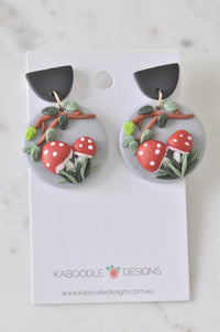 Pair of clay toadstool drop earrings with branch and leaf detail on grey discs, front view on card