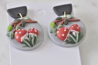 Clay toadstool drop earrings laid flat showing grass, leaves and branch detail