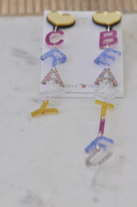 Both letter drop earrings shown flat on marble surface with Kaboodle Designs branded card behind