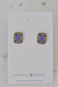 Pair of enamel stud earrings shaped like a yellow ornate frame with purple door centre, on backing card