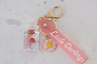 Bottle keychain lying flat beside pink daisy charm and pink Little Darlings strap tag
