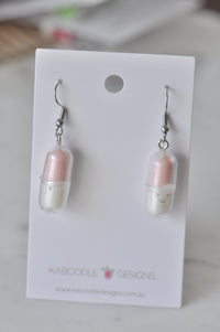Pair of pastel pink and white capsule-shaped dangle earrings with smiling faces, on card