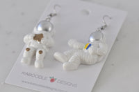 Pair of astronaut drop earrings viewed from above showing silver helmets and star and badge details