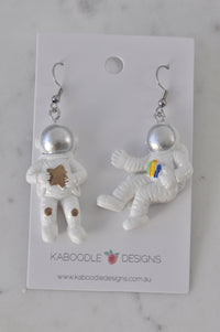 Two resin astronaut earrings on a Kaboodle Designs card, one holding a gold star, one with a coloured badge
