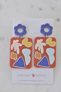 Pair of coral resin drop earrings with abstract cream, mustard and blue shapes on periwinkle flower studs, on card