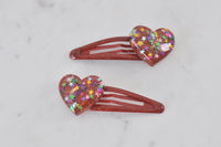 Pair of red resin heart hair clips with rainbow star glitter, laid flat on marble surface