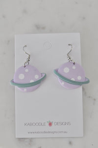 Pair of lilac resin Saturn planet earrings with white dots and sage green ring, shown on jewellery card