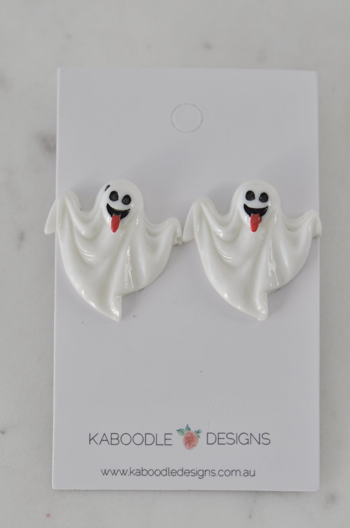 Pair of white ghost stud earrings with smiling tongue-out faces on a display card