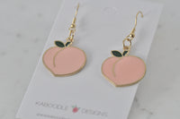 Close-up angled view of two pink peach earrings with green leaf detail on card