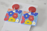 Close-up of two floral resin earrings showing blue, red and yellow flower design