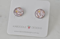 Round stud earrings with pink and yellow floral artwork under glossy resin, shown on jewellery card