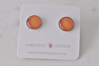 Round handmade stud earrings with warm orange-toned artwork under glossy clear resin