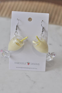 Pair of banana milkshake earrings on card, clear footed glasses with yellow shake, straws and banana slice garnish
