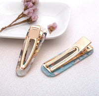Two marbled acrylic barrette clips, one grey and white, one blue, shown open beside dried flowers