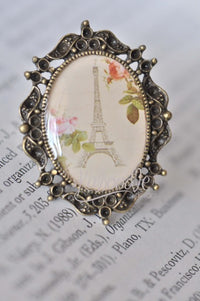 Vintage-style ring with resin Eiffel Tower and floral scene in ornate bronze frame, close-up front view