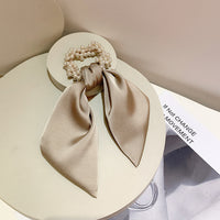 Beige fabric scrunchie covered in clustered faux pearls with a satin bow tail, on a round stand