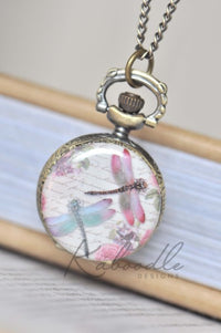 Dragonfly pocket watch necklace with watercolour design resting against a book