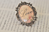 Oval vintage-style ring with 'Love' script over floral background in ornate bronze filigree frame