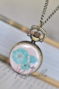 Pocket watch necklace with blue poppy flowers and bird design on pink background